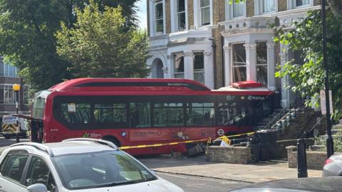 A red bus is seen crashed into a building, with a taped off cordon surrounding it.