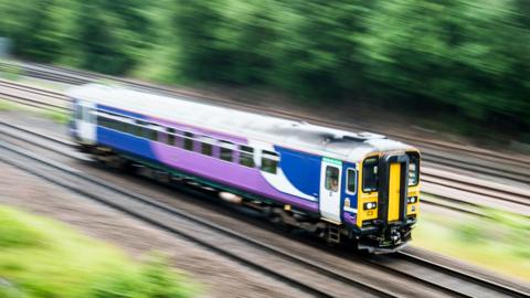 A blue, white and purple train, part of the Northern Trains service, pictured mid-journey.