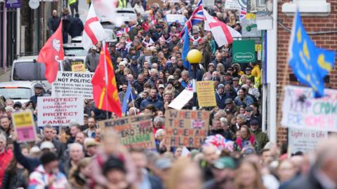 People walking down a street carrying signs and flags protesting about plans to house asylum seekers at a former military site in Crowborough