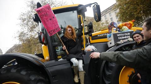 a woman sits on the side of a tractor holding a sign with a drawing of a cow that reads "moooove over starmer" at a protest in london