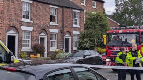 Police officers and a fire engine on a residential street next to a house which has been cordoned off. Several other vehicles are parked on the road.