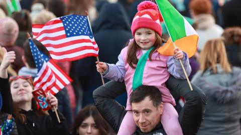 A young girl holds US and Irish flags as she sits on her father's shoulders while they wait for Joe Biden in Ballina