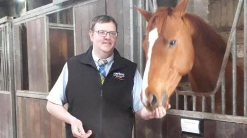 A man with short dark hair and glasses in a black, sleeveless, zip-up top with a short-sleeved shirt underneath and a tartan tie is feeding a chestnut horse with its head sticking out over the stable door 