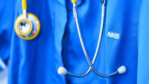 A close-up of someone in a blue NHS shirt. They have a stethoscope around their neck.
