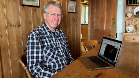 Adrian Corkill, who has grey hair and glasses and is wearing a black and white tartan shirt, sitting in front of a laptop at a wooden table.