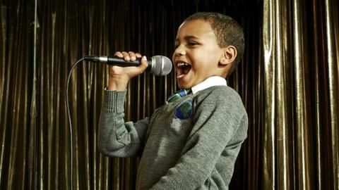 A young boy in his school uniform singing into a microphone