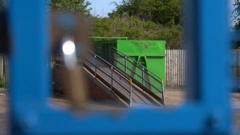 A green skip at a household recycling centre viewed through blue railings