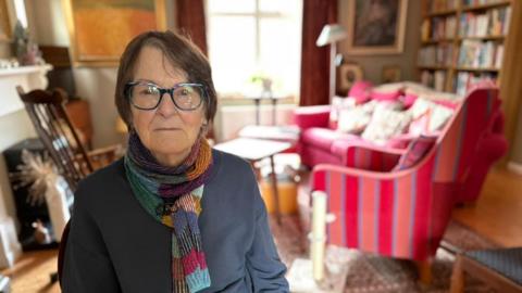 A woman is in her front room, looking at the camera. She is wearing a dark grey top with a multi-coloured scarf. She has short brown hair and glasses.
