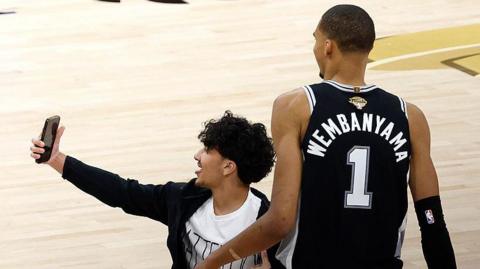A fan runs on to the court and takes a photo with Victor Wembanyama during game one of the 2026 NBA Finals