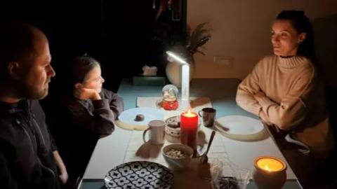 A quiet, candlelit dinner scene with four people gathered around a rectangular table. The only sources of light are a few candles and a small lamp in the center, casting a warm, intimate glow over the setting. The table is modestly set with mugs, bowls of food, a teapot, and a snow globe.