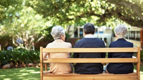 Three older people, seen from behind, sat side-by-side on a bench on a sunny day