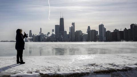 A woman takes a picture as steam rises from the city buildings and Lake Michigan in Chicago