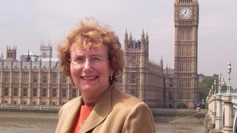 An older picture of Dame Annette standing in front of the Houses of Parliament. She has red hair and wears a beige coat and orange top.