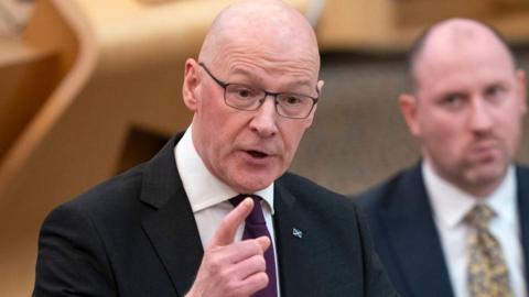 First Minister John Swinney gesticulates with his left index finger with Health Secretary Neil Gray sitting in the background