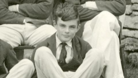 Black and white photo of a young Alan Turing. He is dressed in a shirt, tie and blazer.