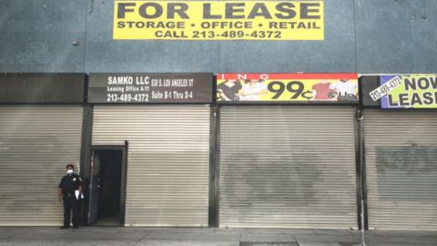 A security guard wears a face mask while standing outside shuttered shops and a For Lease sign amid the global coronavirus pandemic on 30 March 2020 in Los Angeles, California