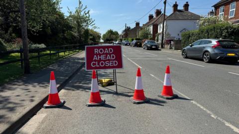 Badshot Lea Road near Farnham. A red sign says 'ROAD AHEAD CLOSED' in white letters. There are four red and white traffic cones in front of it. There are trees and plants on one side of the road, on the other are houses and cars.
