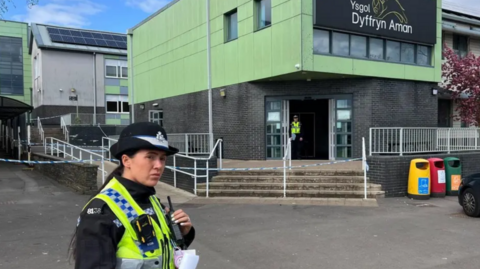 A female police officer in uniform stood outside Ysgol Dyffryn Aman which is a green and grey building with a police cordon in front of it.