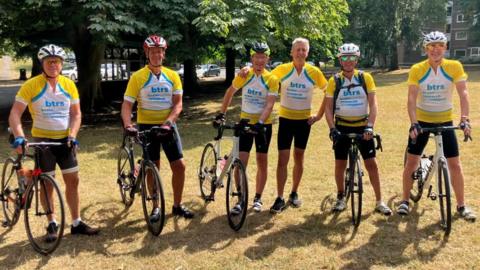 Six men wearing yellow branded cycling jerseys which read "B T R S, brain tumour research and support". They are standing with bicycles in a park, and most wear cycling helmets.
