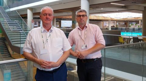 A man with white hair, a short sleeved white shirt, navy blue trousers and a white and green lanyard leans on a railing with one elbow. His hands are linked, and his facial expression is neutral. He is beside another male, who has grey hair, is wearing glasses and a pink shirt with his sleeves rolled up to his elbows. He's wearing black trousers and a white lanyard. The second male is also leaning on a railing with one elbow, and also has a neutral expression with his hands linked. There are flights of stairs in the background.