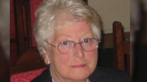 Janet Purkess is seated in a room. She has neat, wavy, short, silver hair and glasses.