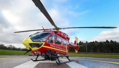 A red and yellow helicopter on a grey and white helipad on the ground of a green field. There are trees in the background.