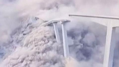Bridge collapses leaving clouds of white smoke and dust, part of the remaining bridge is on the right of the image while the collapsed part of the bridge is on the left of frame.