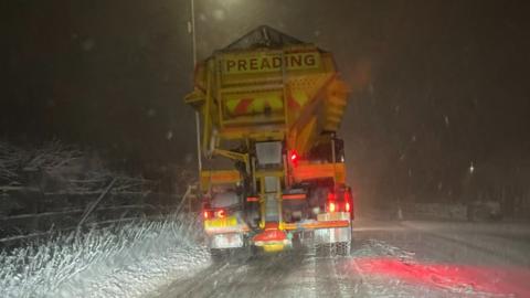 A gritter heads along a snow covered road at night