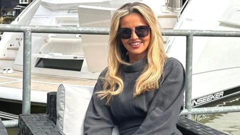 A young white woman with long blonde hair sits in front of a yacht. She's on an outdoor armchair, wearing sunglasses and a loose, polo-necked tracksuit in a luxurious dark grey colour.