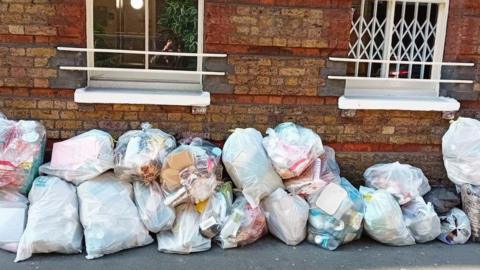 A row of clear plastic rubbish bags stacked up and filled with mixed household waste, including cardboard boxes, food containers, and plastic bottles, lined up on a pavement in front of a brick building with two white-framed windows and metal security bars.