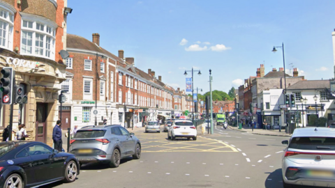 Epsom High Street at the junction with Ashley Road has shops on either side and people walking on the pavements. Cars are using the junction and there are lampposts and traffic lights.