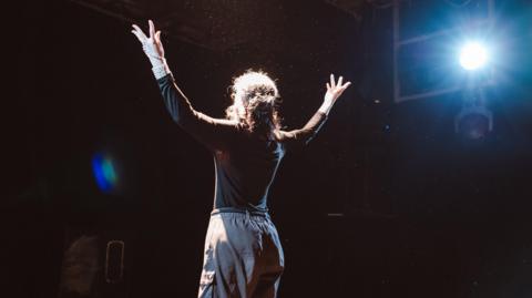 A performer on stage dressed in a black top and grey trousers. 
A spotlight can be seen at the side of the stage.