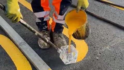 Yellow liquid is poured into a tool which spreads it onto the road surface