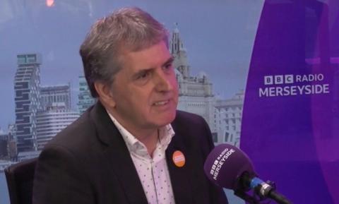 Steve Rotheram, wearing a black jacket and white patterned shirt, sits at a purple microphone in BBC Radio Merseyside's studio