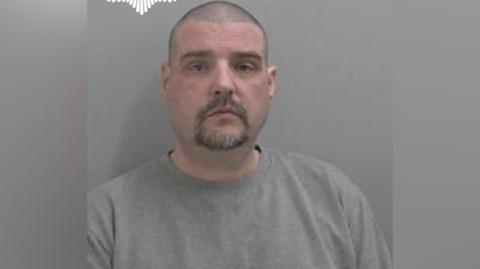 Chris Hemming pictured in a mugshot. He has a blank expression and wears a grey jumper. He has dark stubbly hair and a dark beard. 