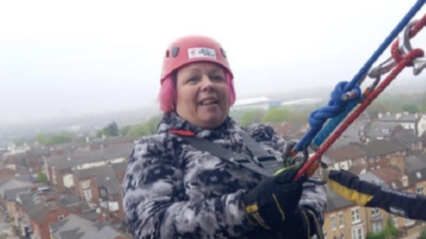 A woman wearing a pink helmet, a coat and a harness holds on to two ropes as she abseils down a building, with houses, streets and parked cars visible below her.