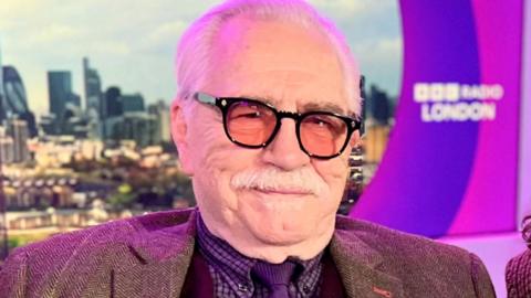 Actor Brian Cox, a white man with white hair, black glasses and a moustache smiles at the camera