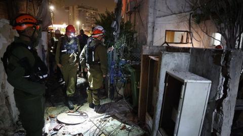 Crews check damage of an Iranian building after strikes. They are wearing hard-hats and you can see exposed wires and brick