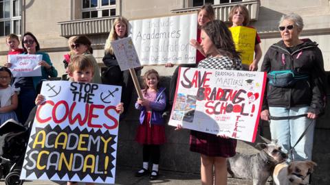 Parents and children stood on pavement outside Isle of Wight council holding banners saying "Stop the Cowes Academy scandal" and "Save Brading school"