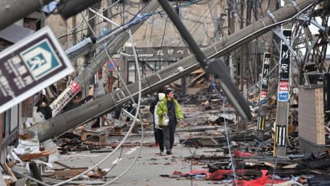People walk past fallen utility poles and damaged buildings in the city of Wajima.