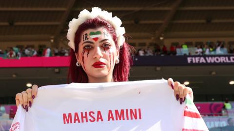 A female fan in the crowd for Wales v Iran holds up a football shirt with "Mahsa Amini - 22" printed on it
