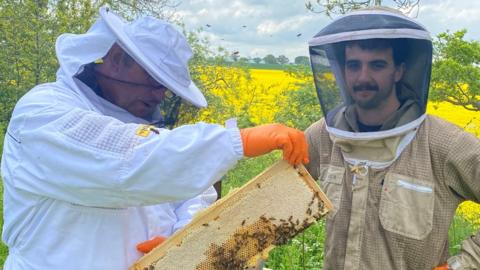 Gruffudd Tomos in a beekeepers suit standing next to Russell Jones, also in a beekeepers suit and hold a hive