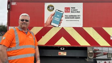 Mr Byers stands in front of the back of a bright red truck with the message wearing high-vis gear