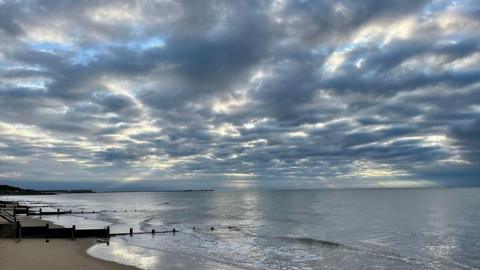 The sky over the sea is a mix of blue, grey and white cloud as the sun rising behind softly glows between them