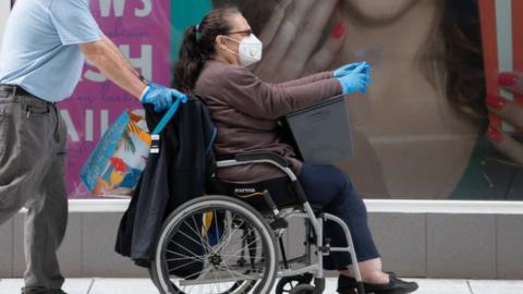 A woman sitting in a wheelchair wearing a mask and gloves, being pushed by a man wearing the same