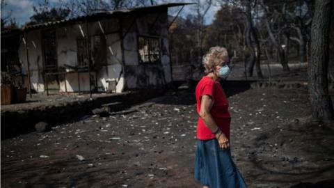 A woman wearing a mask walks in front of her burnt house following a wildfire at the village of Neos Voutzas, near Athens (26 July)