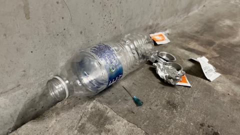 An empty water bottle surrounded by wrappers, dirty tea spoons and syringes
