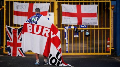 A Bury FC fan, wrapped in an England flag with the word 'Bury' on it, stands outside the locked gates to Gigg Lane in 2019