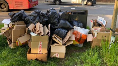 Dozens of broken down boxes and multiple black sacks are piled up behind a bus stop pole on the edge of Wandsworth Common