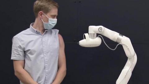 A person wearing a face mask and a rolled-up shirt sleeve stands next to a robot which is about to administer an injection